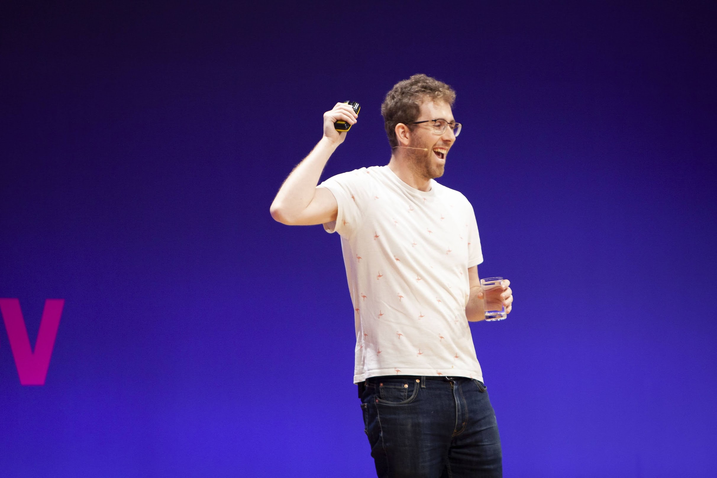 Me laughing and speaking with a conference mic on my face, wearing a white t-shirt with small pink flamingos, holding a cup of water